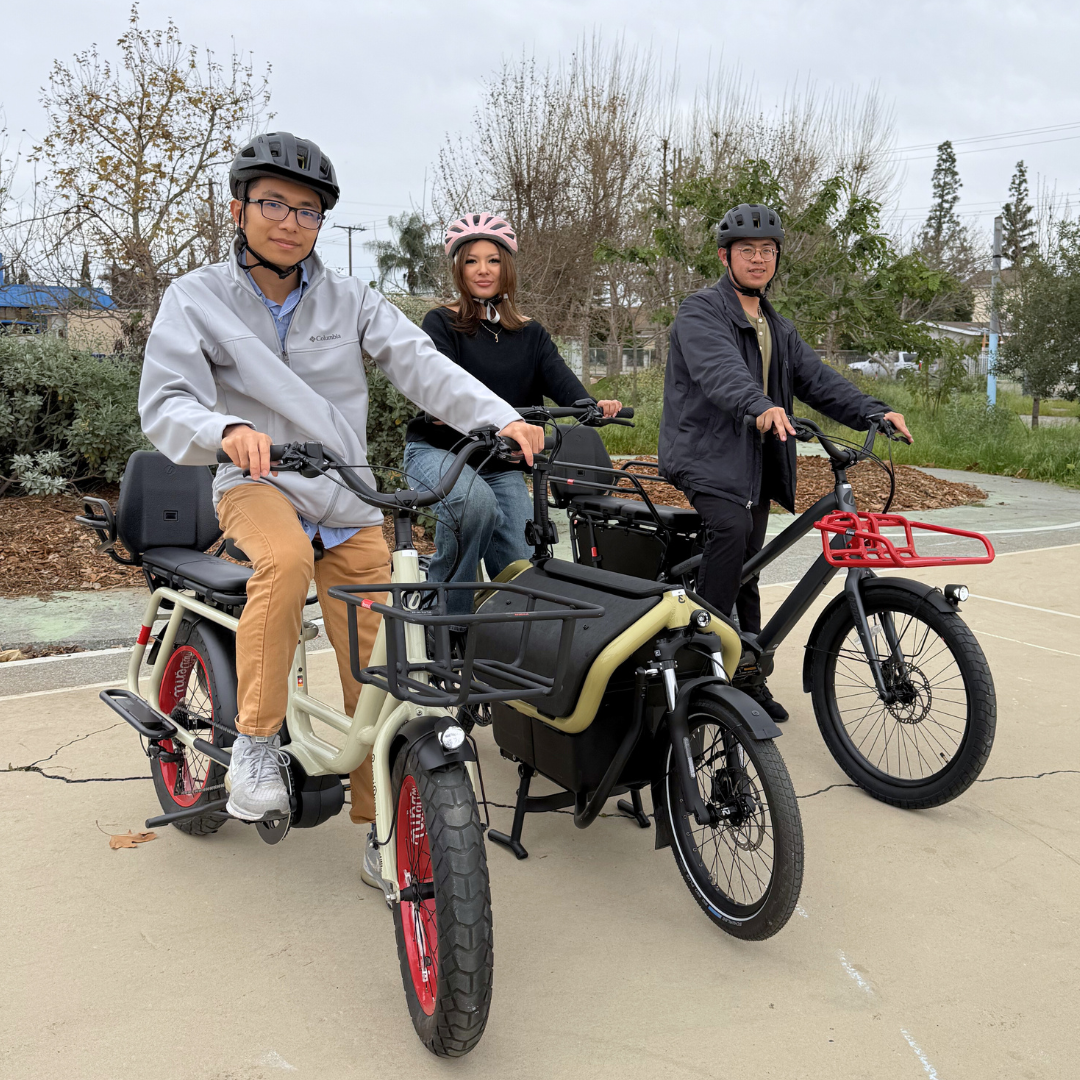 Three people standing with electric cargo bikes on a paved path in a neighborhood.
