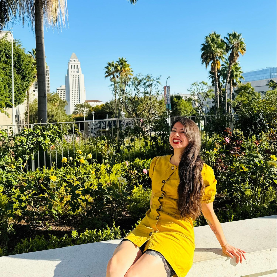 Gabby Magaña sits smiling in a sunny garden space with palm trees and the Los Angeles City Hall building visible in the background. She is wearing a yellow dress surrounded by flowers and greenery.