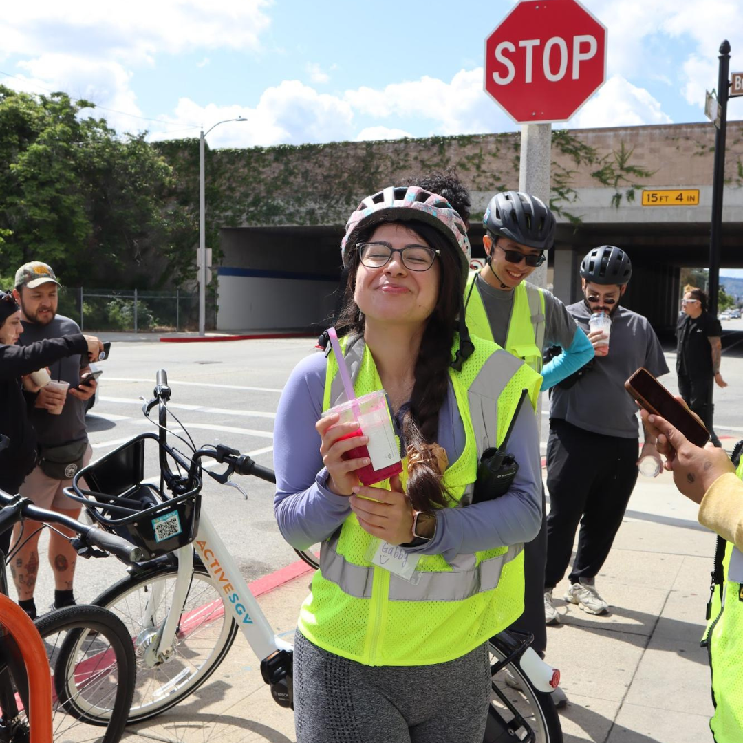 Gabby Magaña stands with a group during a community bike ride, wearing a helmet and reflective safety vest while holding a drink near parked bicycles and a city street intersection.