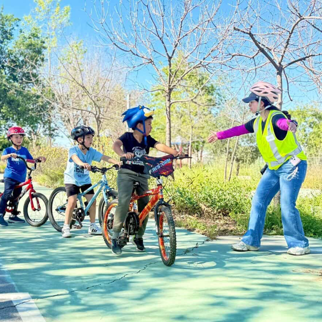 Gabby Magaña helps lead a children’s bike activity outdoors. She wears a safety vest and helmet while guiding young riders on a paved path lined with trees.