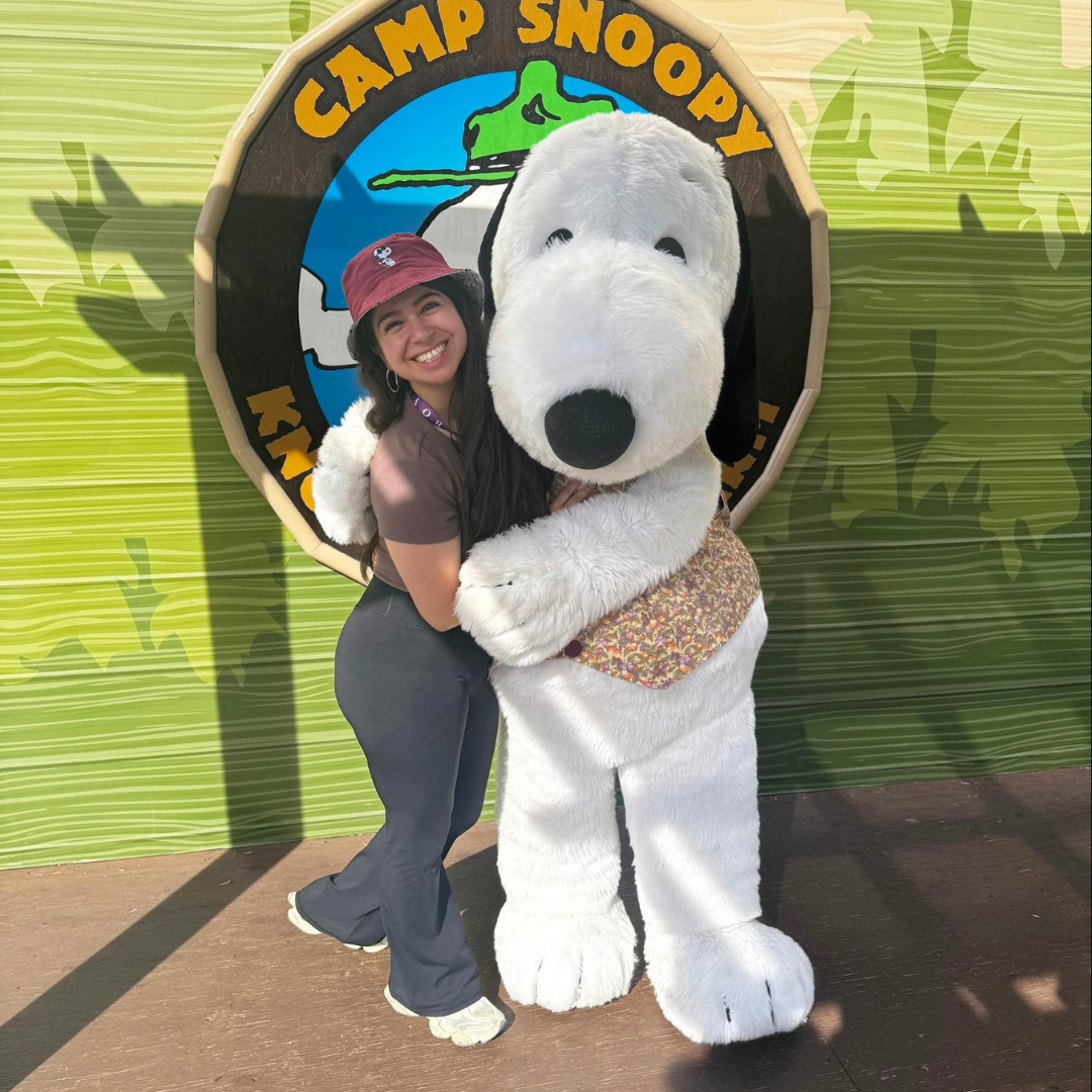 Gabby Magaña smiles while posing with the Snoopy character at Camp Snoopy. She wears a bucket hat and casual outfit in front of a themed backdrop.