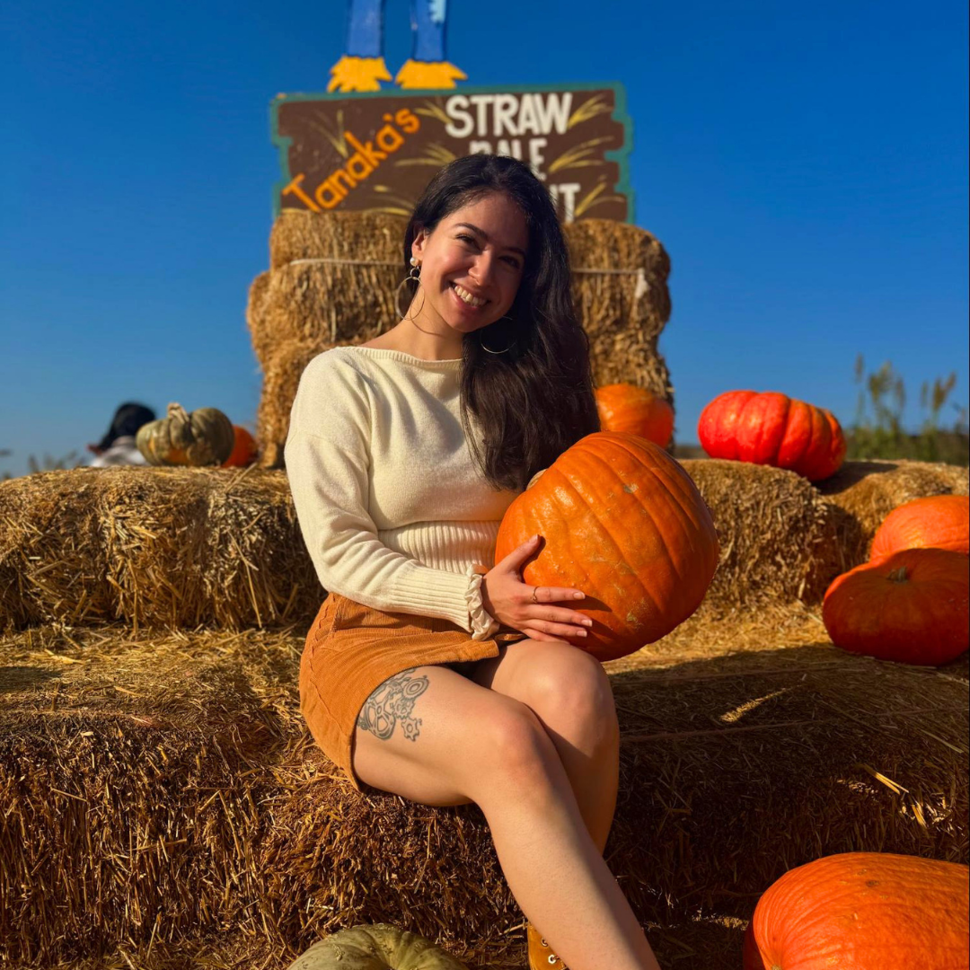 Gabby Magaña smiles while sitting on stacked hay bales at a pumpkin patch, holding a large pumpkin. Orange pumpkins and straw bales surround her under a clear blue sky.