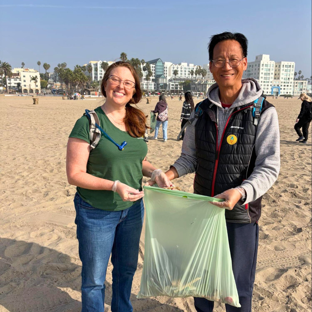 Two participants smiling while holding a bag of collected trash during a beach clean-up.