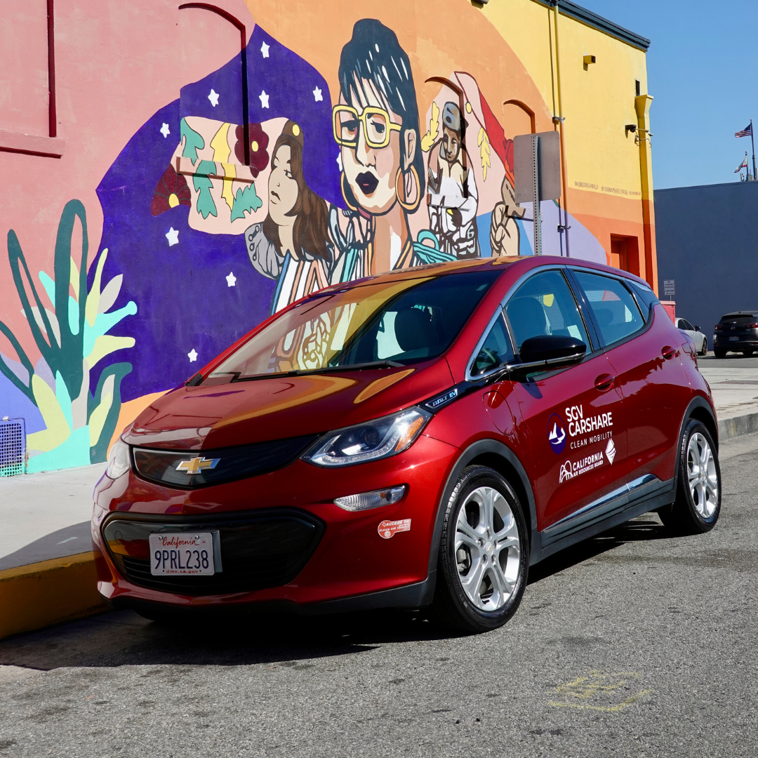 A red SGV Carshare electric vehicle is parked in front of a colorful community mural in El Monte.