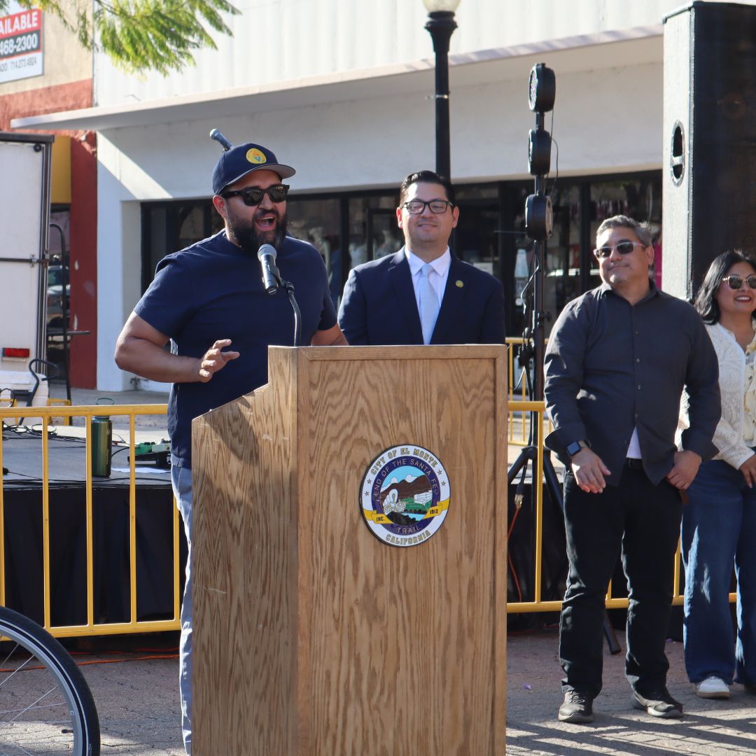 A speaker addresses attendees at an outdoor community event podium in El Monte while local leaders stand nearby.