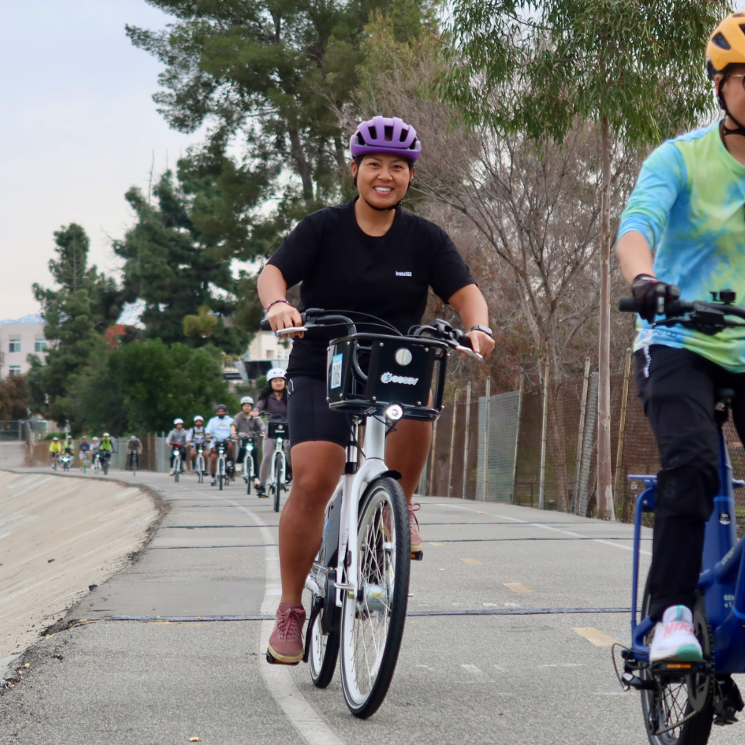 A group of cyclists rides along a paved multi-use trail, led by a rider wearing a purple helmet.