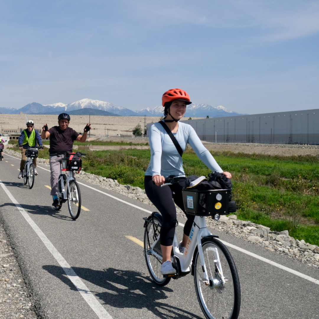 Cyclists ride e-bikes along a paved path with mountain views in the distance.