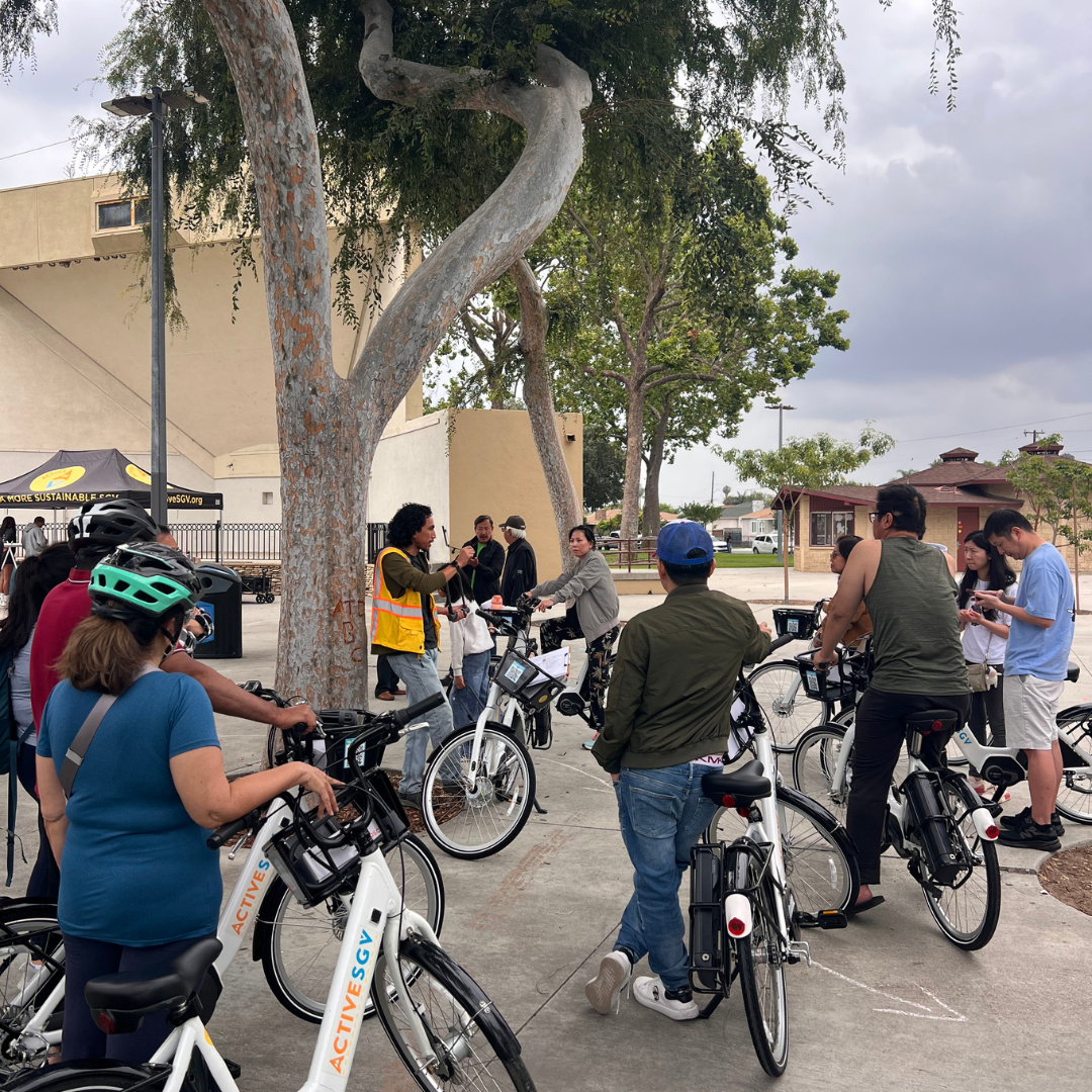 An ActiveSGV ride leader addresses a group of participants gathered with e-bikes near a park pavilion.