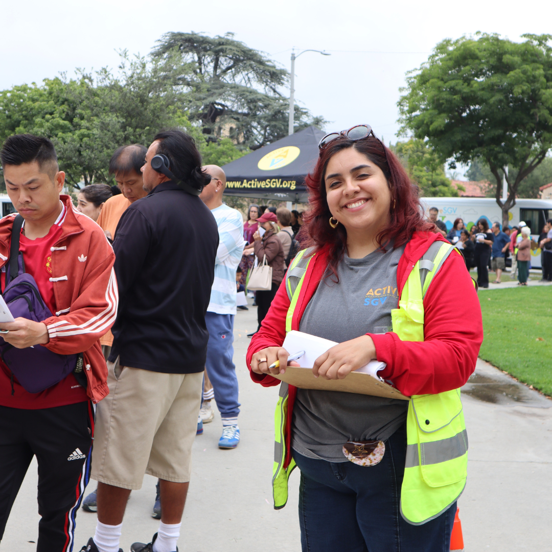 An ActiveSGV volunteer wearing a safety vest smiles while holding a clipboard during event check-in.