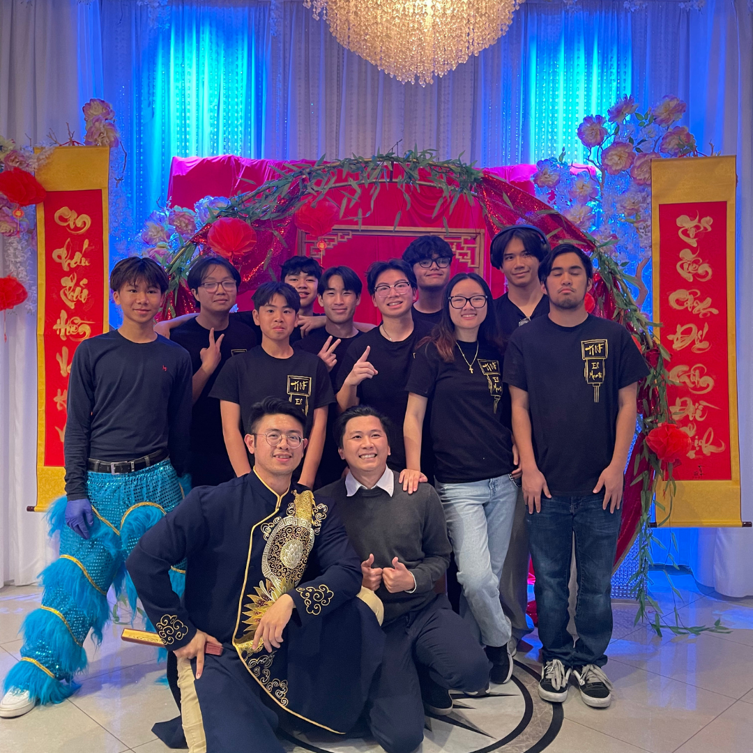 Dat Cao poses with members of a Vietnamese cultural performance group in front of a Lunar New Year themed backdrop decorated with red and gold banners.