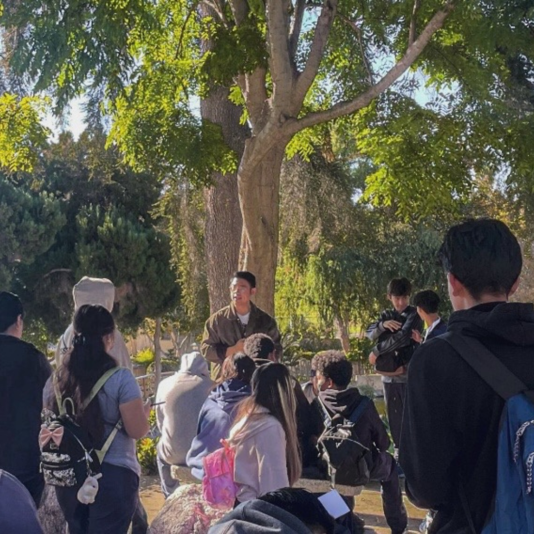 Dat speaks to a group of students gathered outdoors beneath trees during an educational activity.