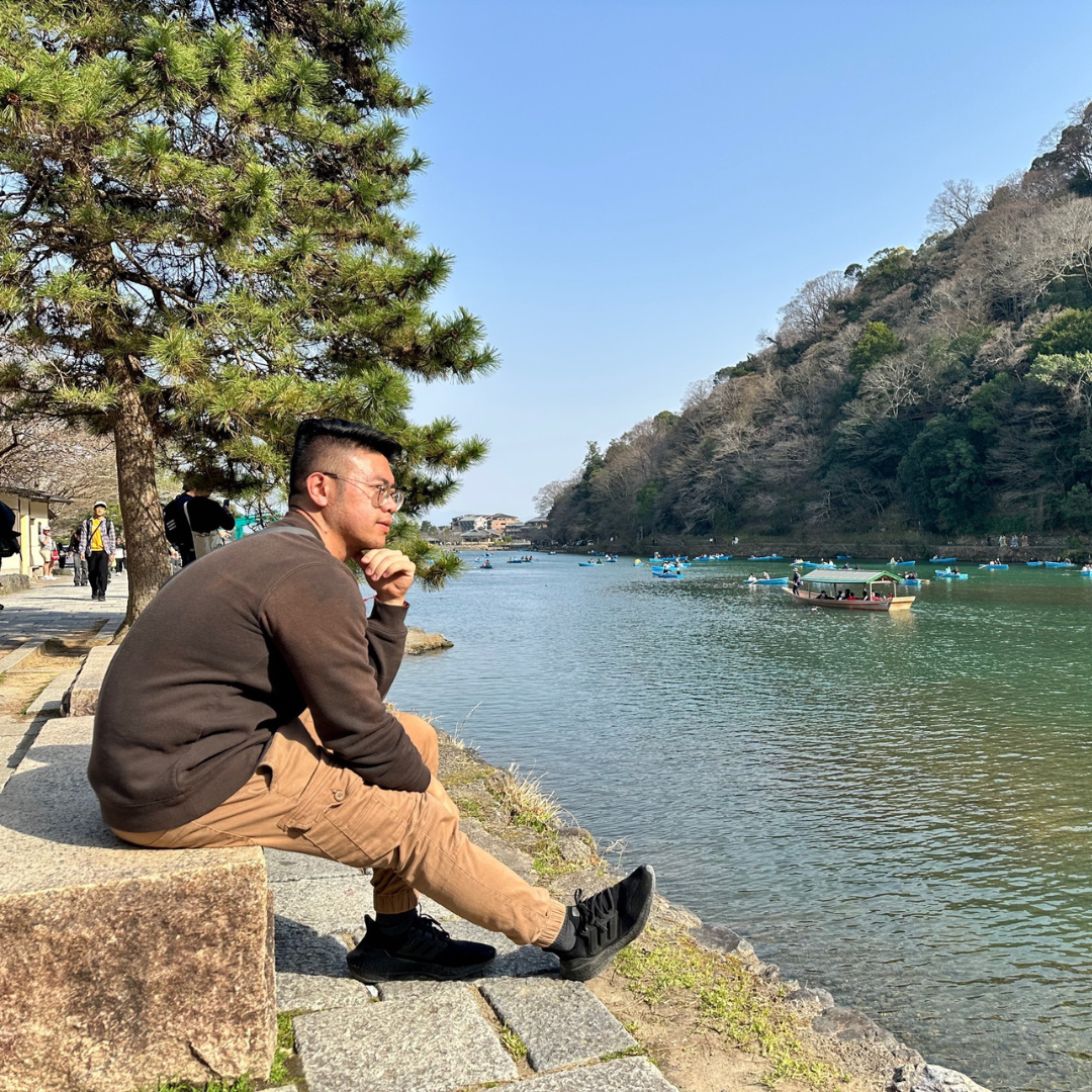 Dat sits along the edge of a river in Japan, looking out over the water and passing boats.