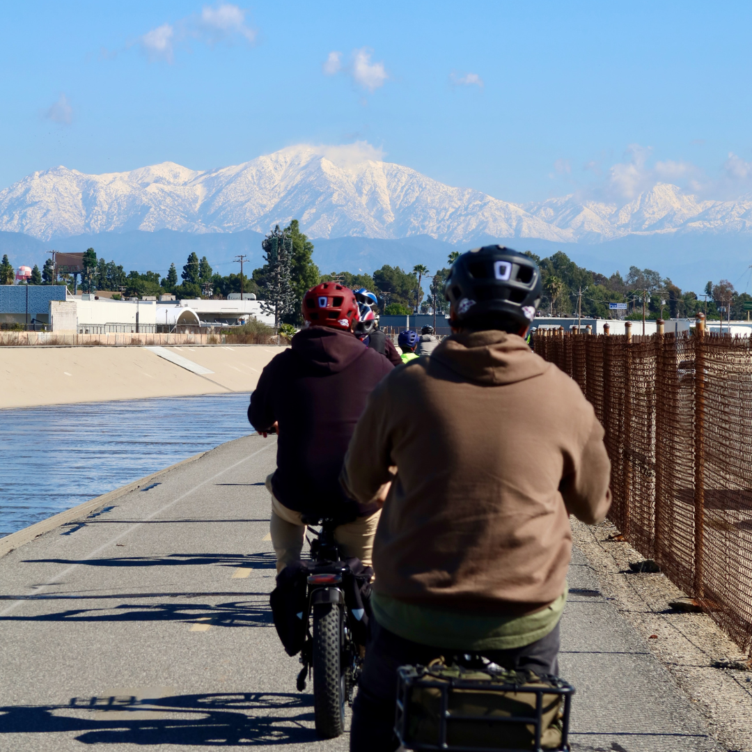 Cyclists ride along a river channel with snow-capped mountains in the distance.