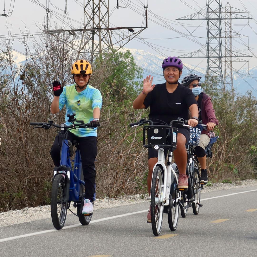 Three cyclists smile and wave while riding along a paved bike path with mountains in the background.