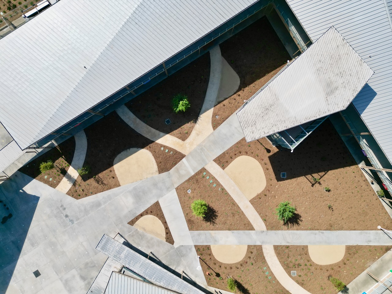 Aerial view of the Shirpser campus courtyard showing curved decomposed granite pathways, newly planted garden beds, and young trees between classroom buildings.