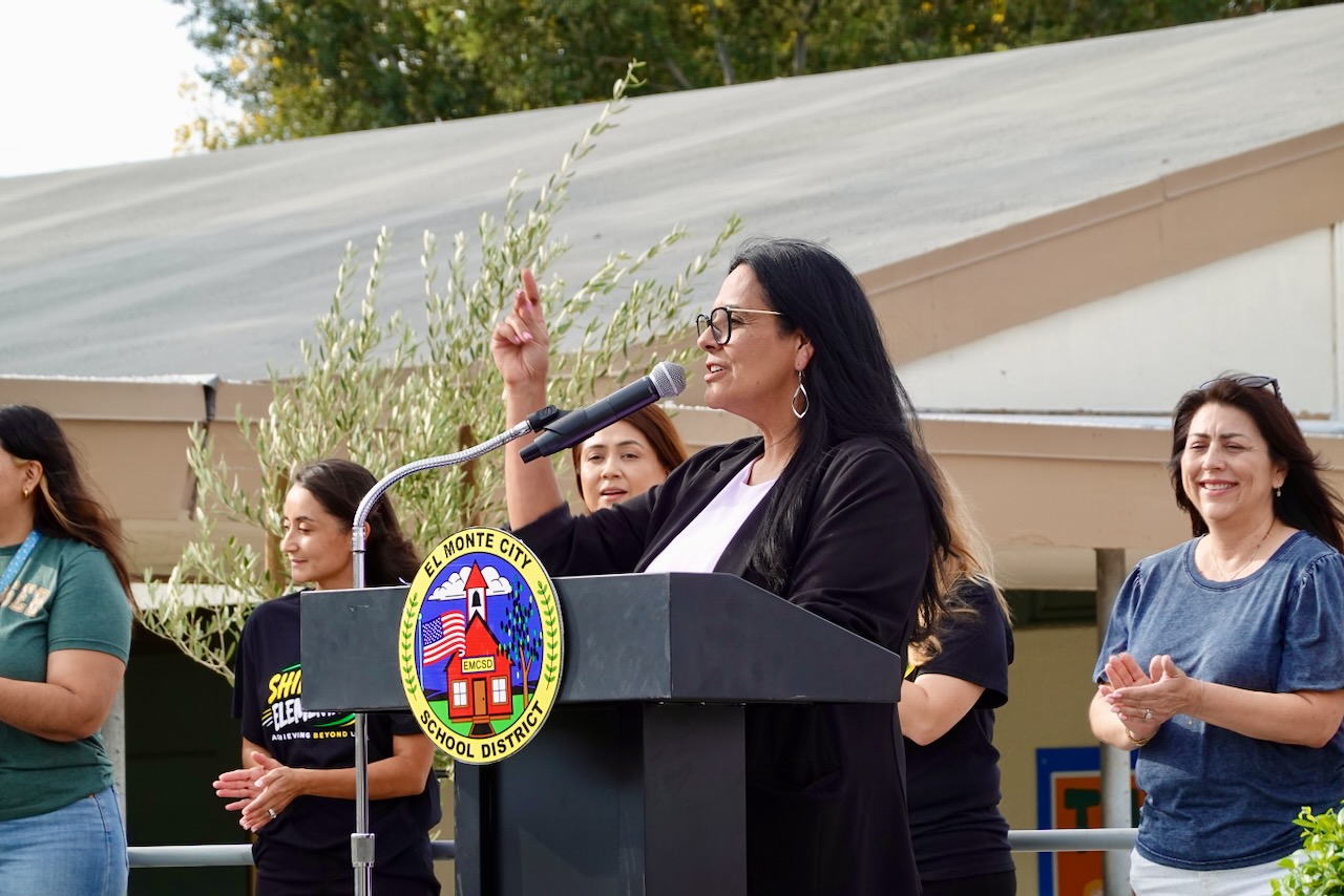 Principal Veronica Ortiz speaks into a microphone at a podium bearing the El Monte City School District seal, with staff applauding beside her.