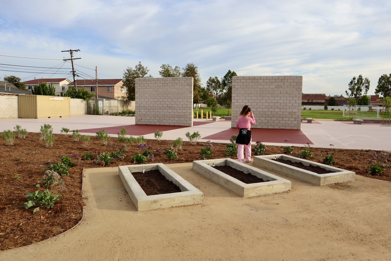 A student walks past three new concrete raised garden beds surrounded by native plants, with handball walls and the play field behind.