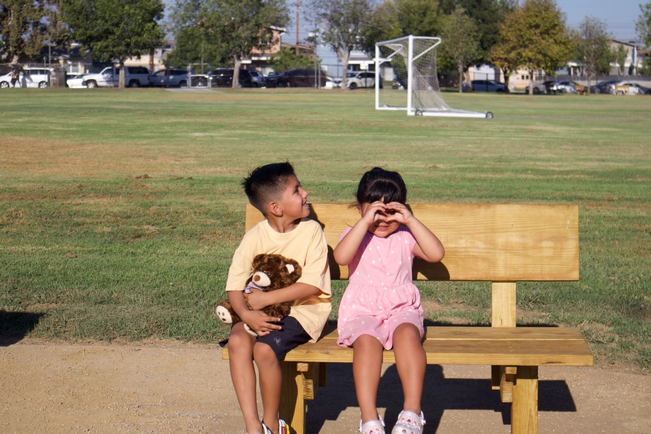 Two young children sit on a new wooden bench beside the green play field at Gidley School, one holding a teddy bear.