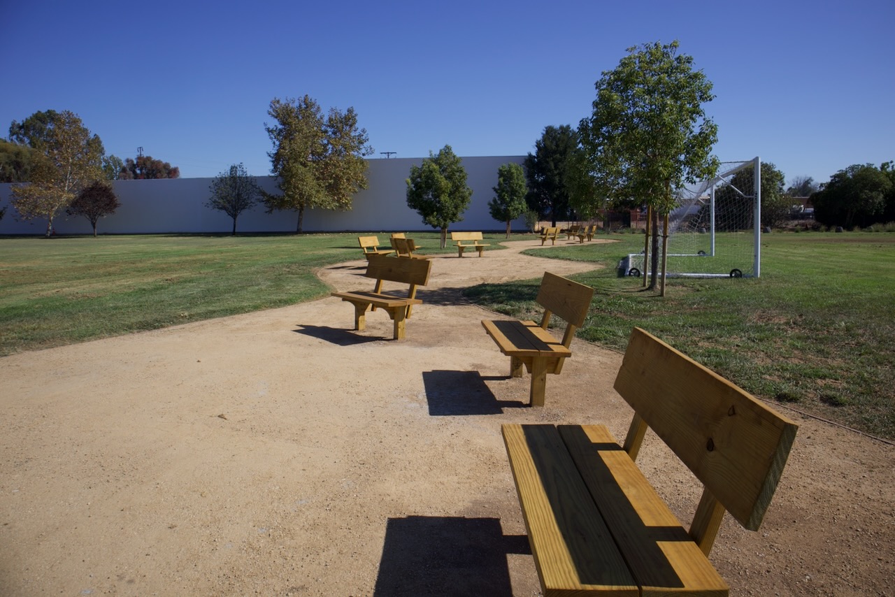 A row of new wooden benches set along a decomposed granite walking path beside the Gidley play field, with young trees behind.
