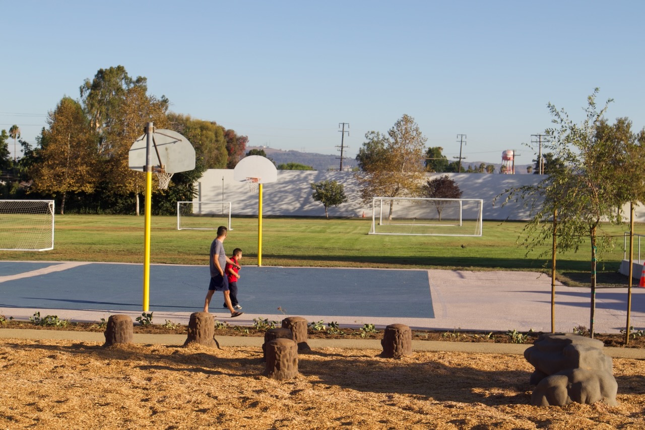 An adult and a young child walk past the resurfaced basketball courts at Gidley, with nature play stepping stones in the foreground.