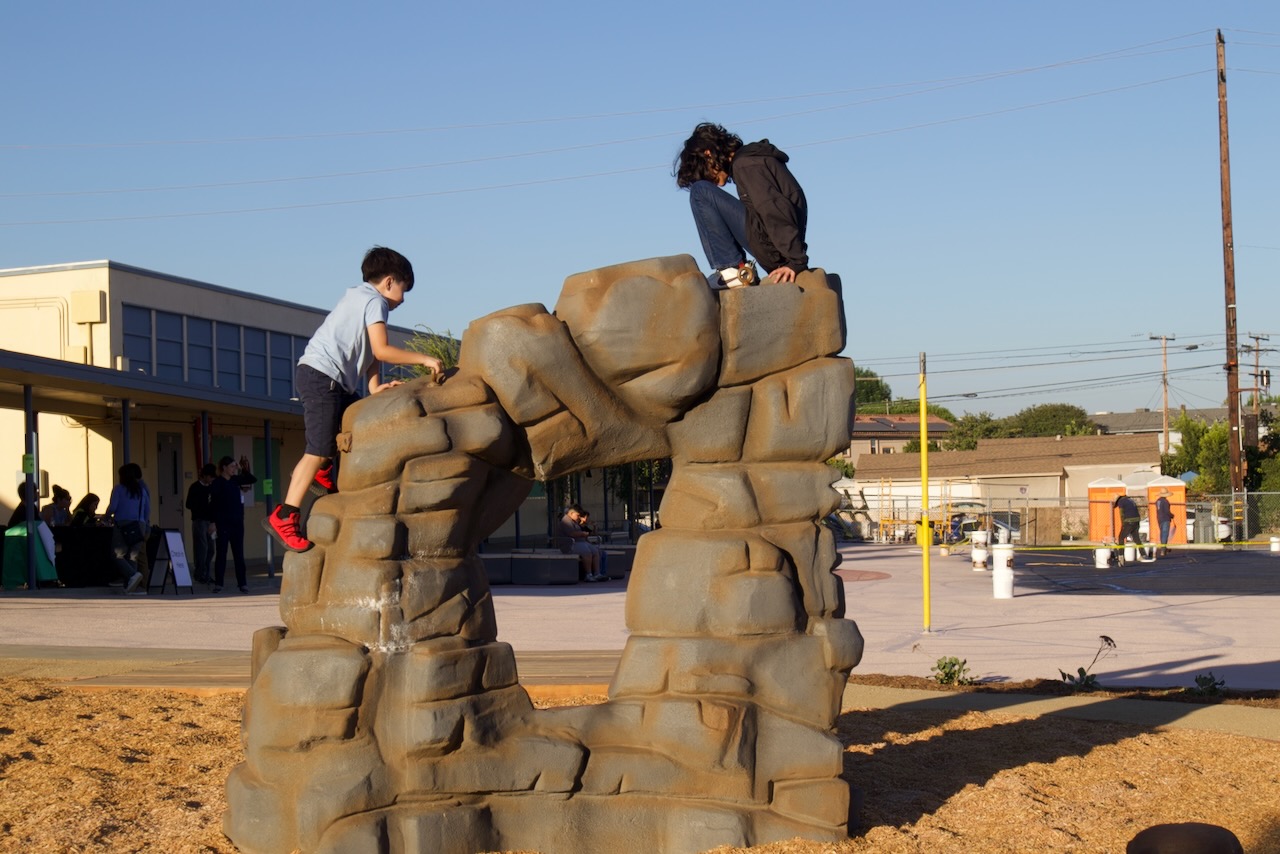 Two students climb a boulder play structure in a mulched play area at Gidley School at sunset.