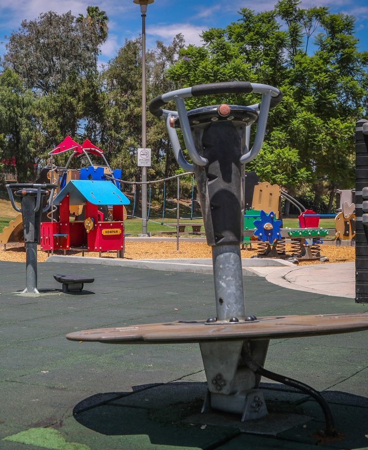 A neighborhood park playground in Avocado Heights with a colorful playhouse and a spinner seat under tall shade trees