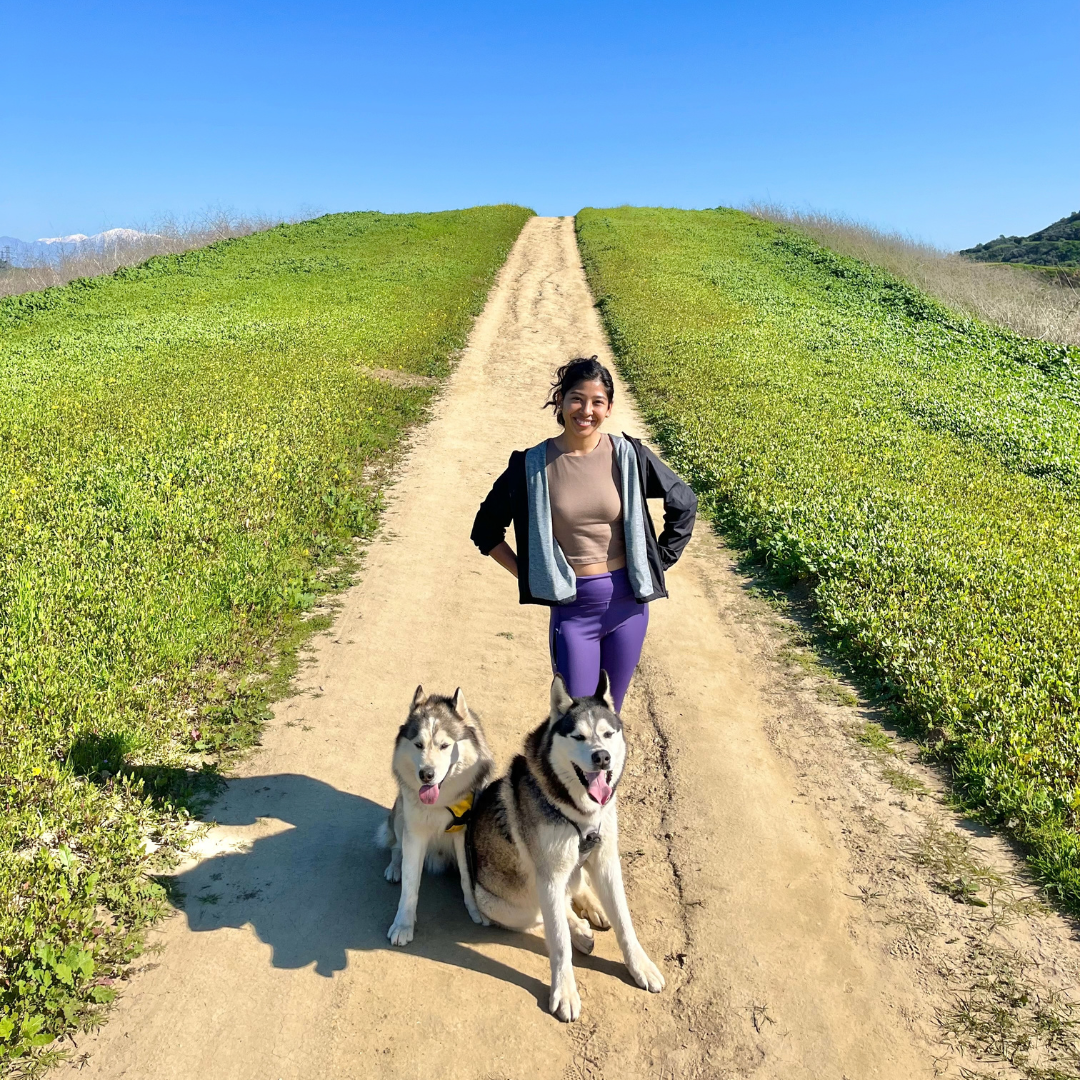 Yvette stands on a dirt trail with two huskies, surrounded by green hills under a clear blue sky.