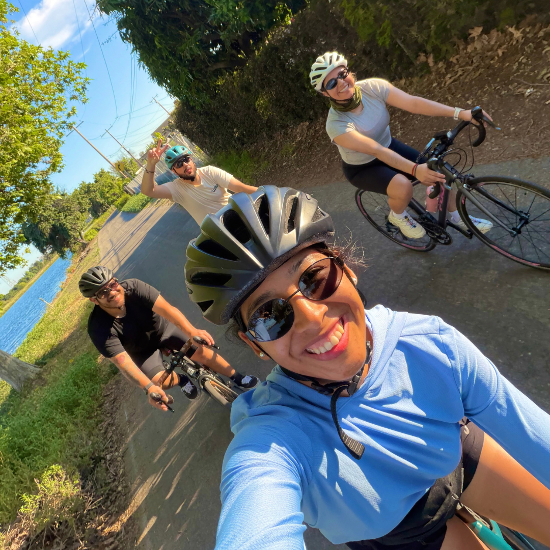 Yvette smiles for a selfie with three other cyclists during a group bike ride along a tree-lined path next to the water.