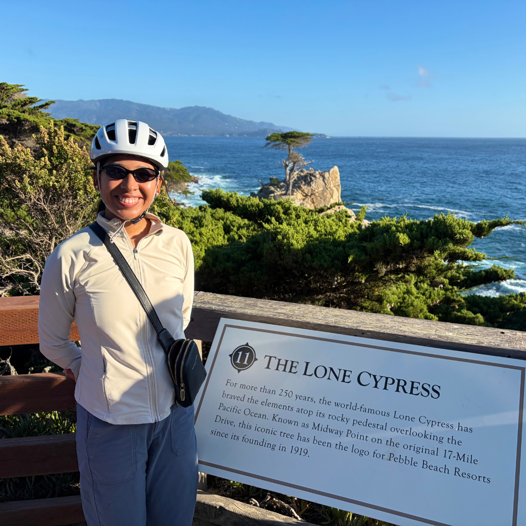 6. Yvette, wearing a bicycle helmet and sunglasses, smiles at a scenic coastal overlook near the Lone Cypress along 17-Mile Drive.