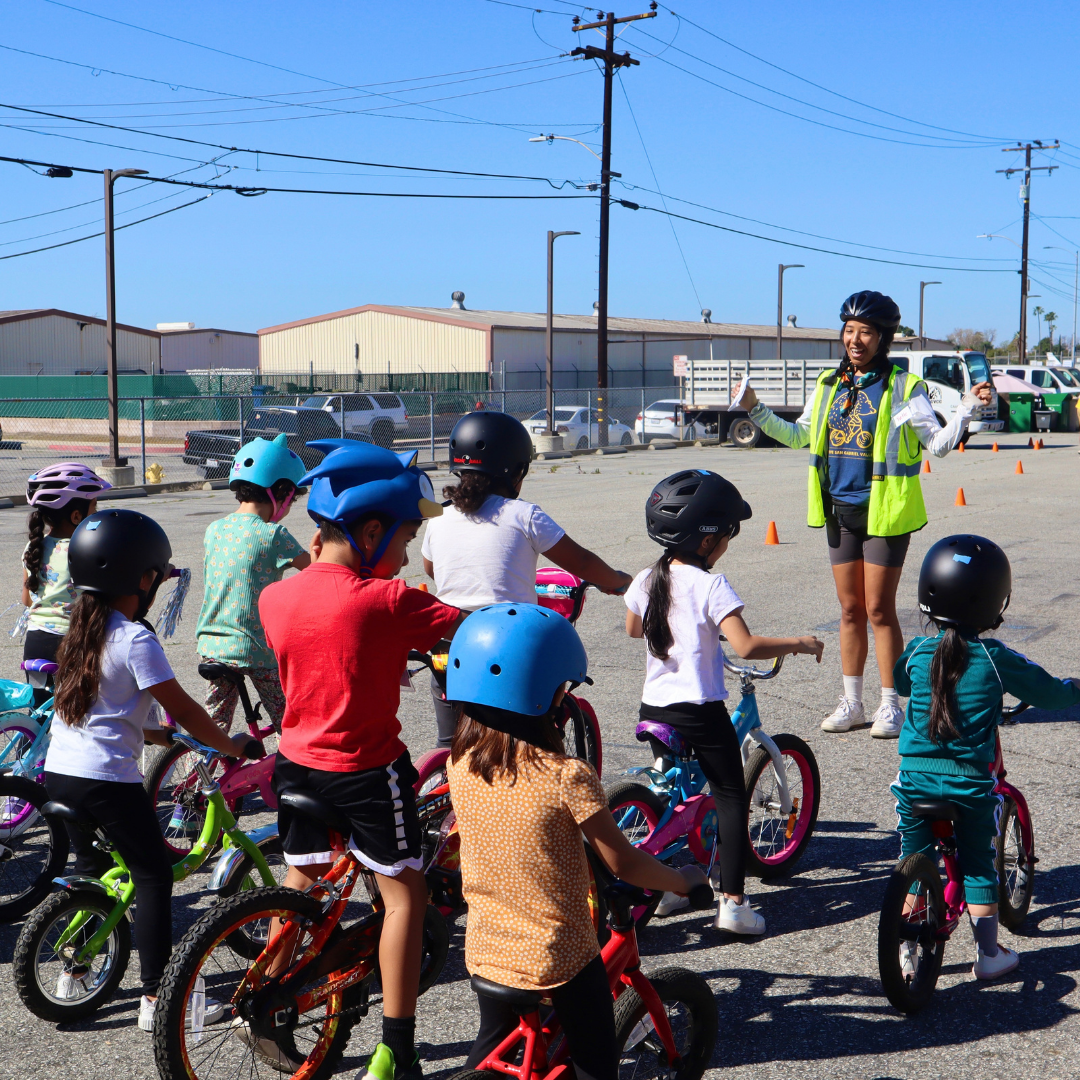 Yvette, wearing a helmet and safety vest, gives instructions to a group of children on bicycles during an outdoor bike rodeo.