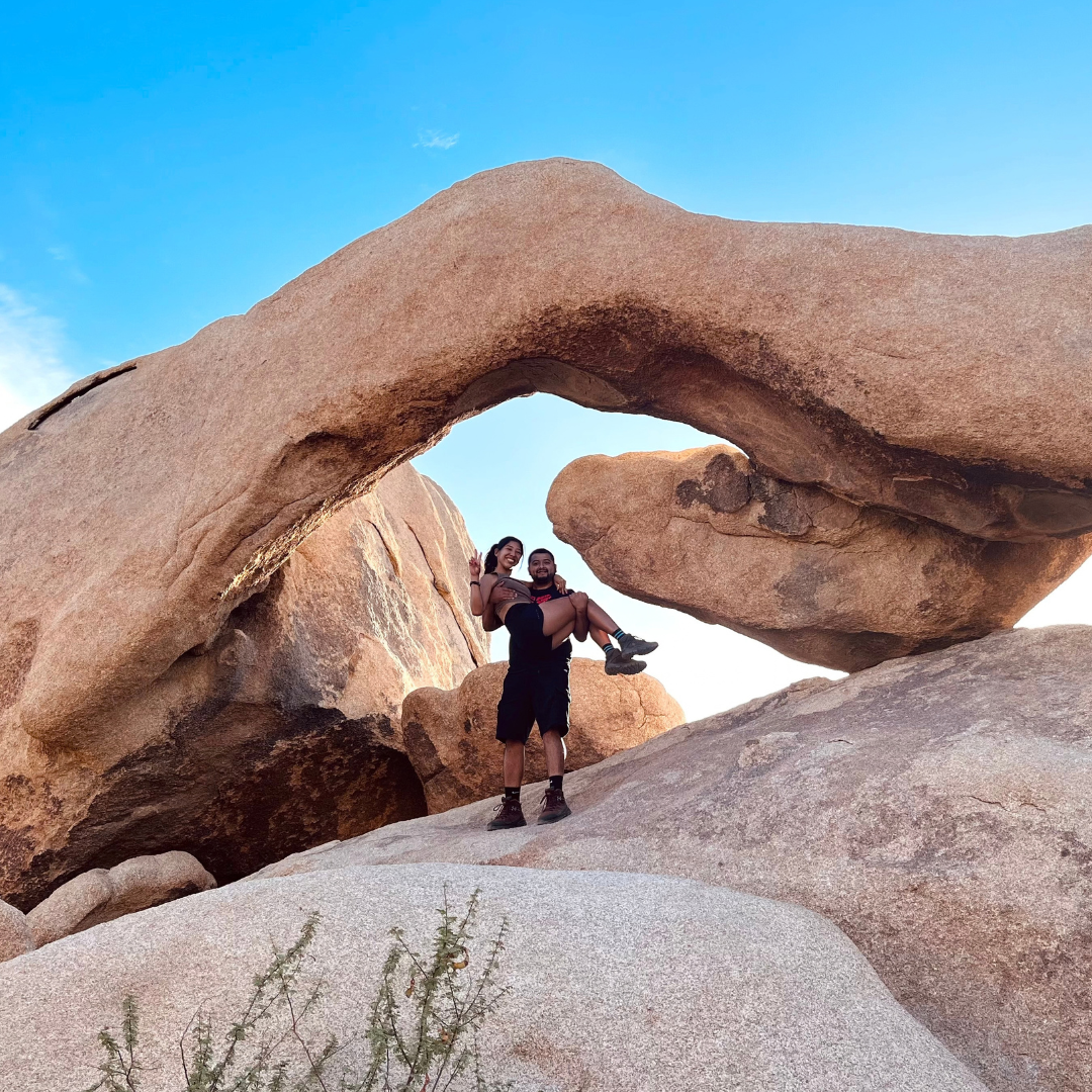 Yvette and her spouse pose beneath a large natural rock arch during a hike in a desert landscape.