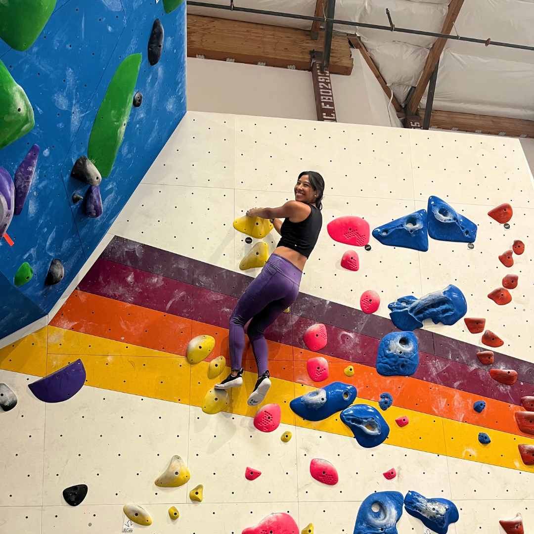 Yvette smiles while climbing an indoor bouldering wall covered with colorful climbing holds.