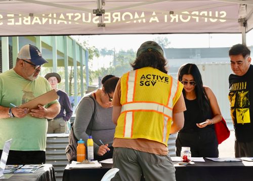 An ActiveSGV team member in a yellow safety vest helps community members sign up at an outdoor event table.