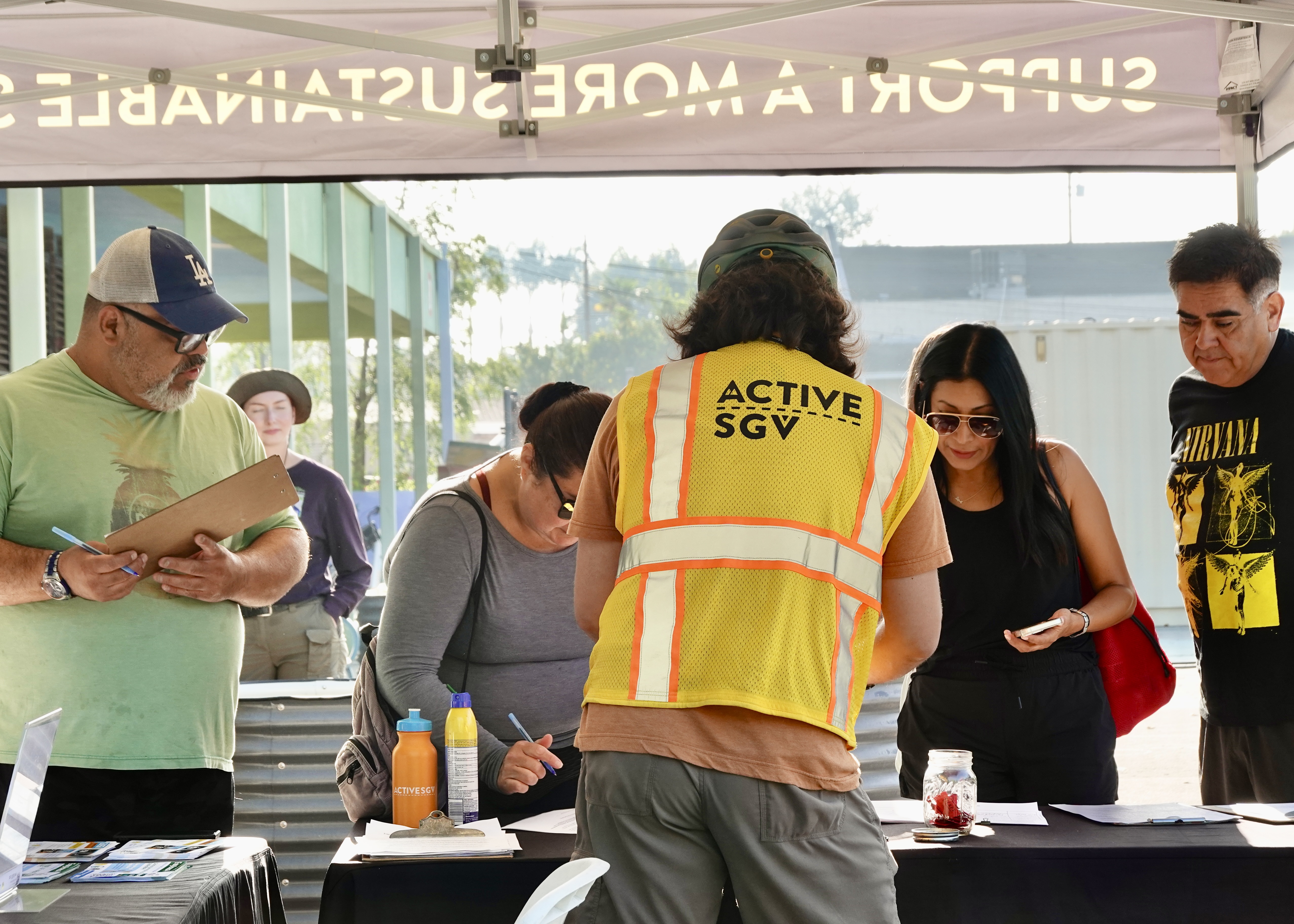 An ActiveSGV team member in a yellow safety vest helps community members sign up at an outdoor event table.