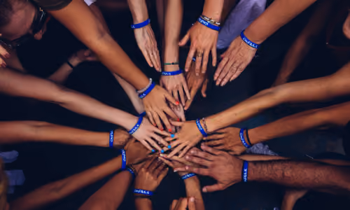 Multiple hands of diverse individuals stacked together in a circle, all wearing blue wristbands symbolizing unity and teamwork.