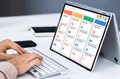 Person typing on a wireless keyboard in front of a tablet displaying a dashboard with multiple colored cards containing icons, graphs, and text, set on a white desk with a phone in the background.