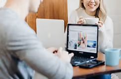 Two people sitting at a table with a laptop open displaying images, one person holding a cup and smiling.