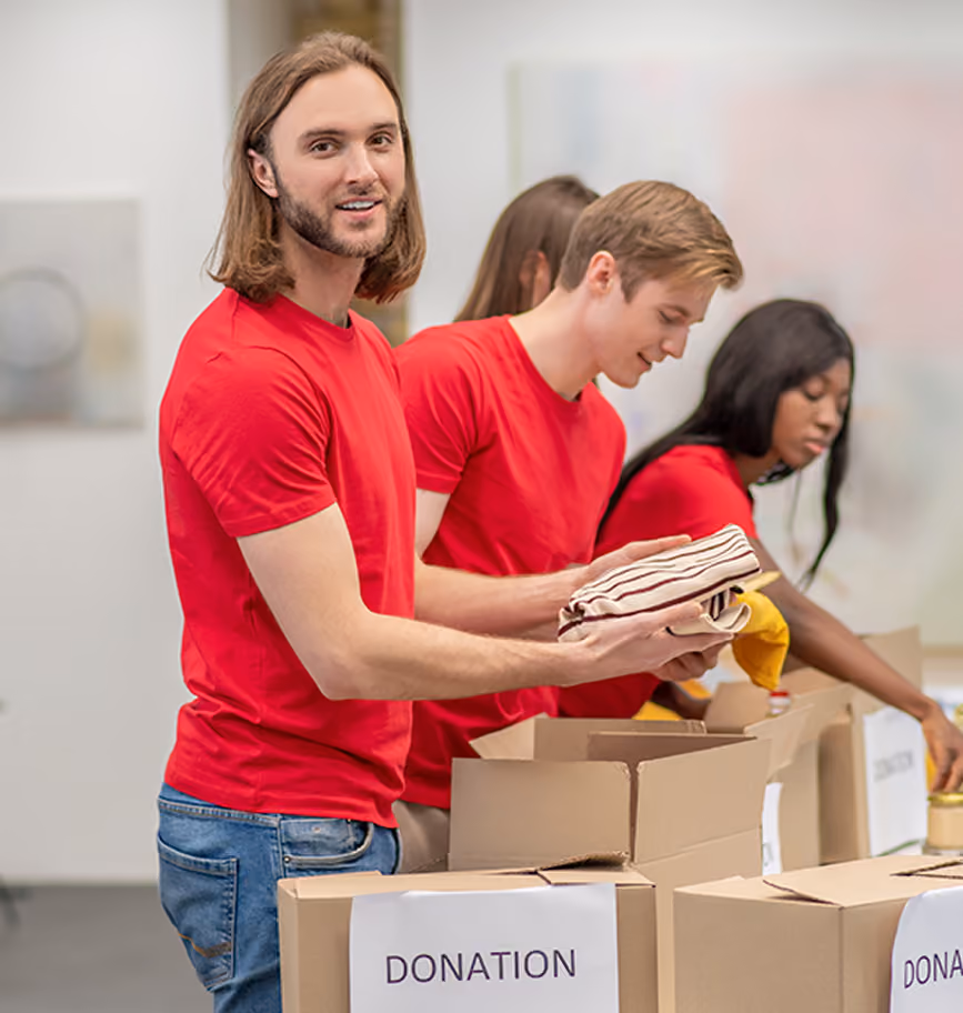 Three young volunteers in red shirts packing items into donation boxes lined up on a table, with one man looking toward the camera holding folded clothes.