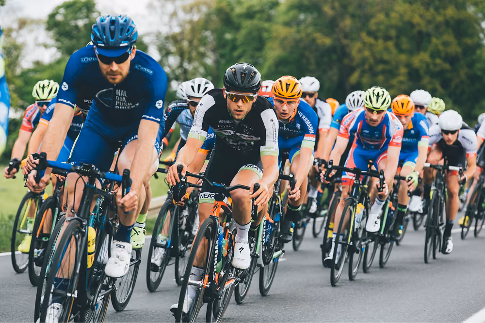 A group of cyclists wearing helmets and colorful racing gear riding closely together on a paved road surrounded by trees, participating in a competitive road cycling race.