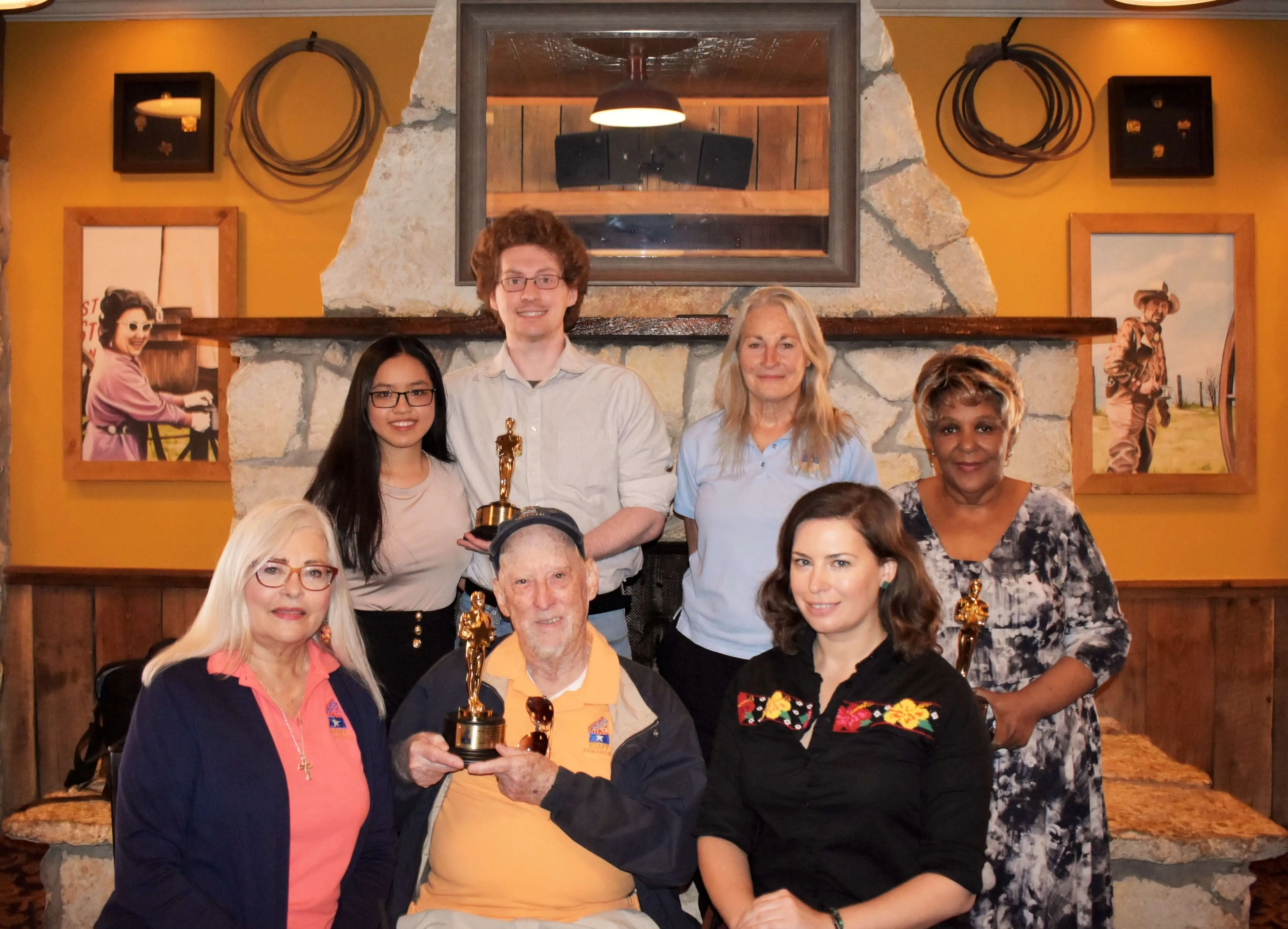 Group portrait of seven people, four standing and three seated, holding Remi Award statuettes, in front of a stone fireplace with a mirror, framed pictures, and coiled cables on yellow walls.