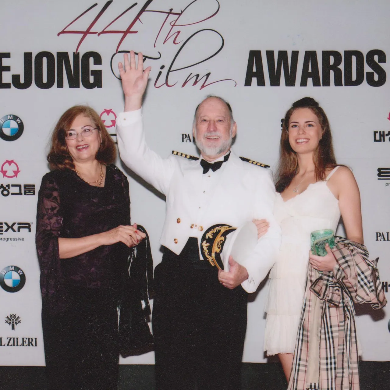 Three people posing at the 44th Blue Dragon Film Awards: a woman in a dark dress on the left, a man in a white formal naval uniform with a black bow tie waving in the middle, and a woman in a white dress holding a plaid coat and a green clutch on the right.