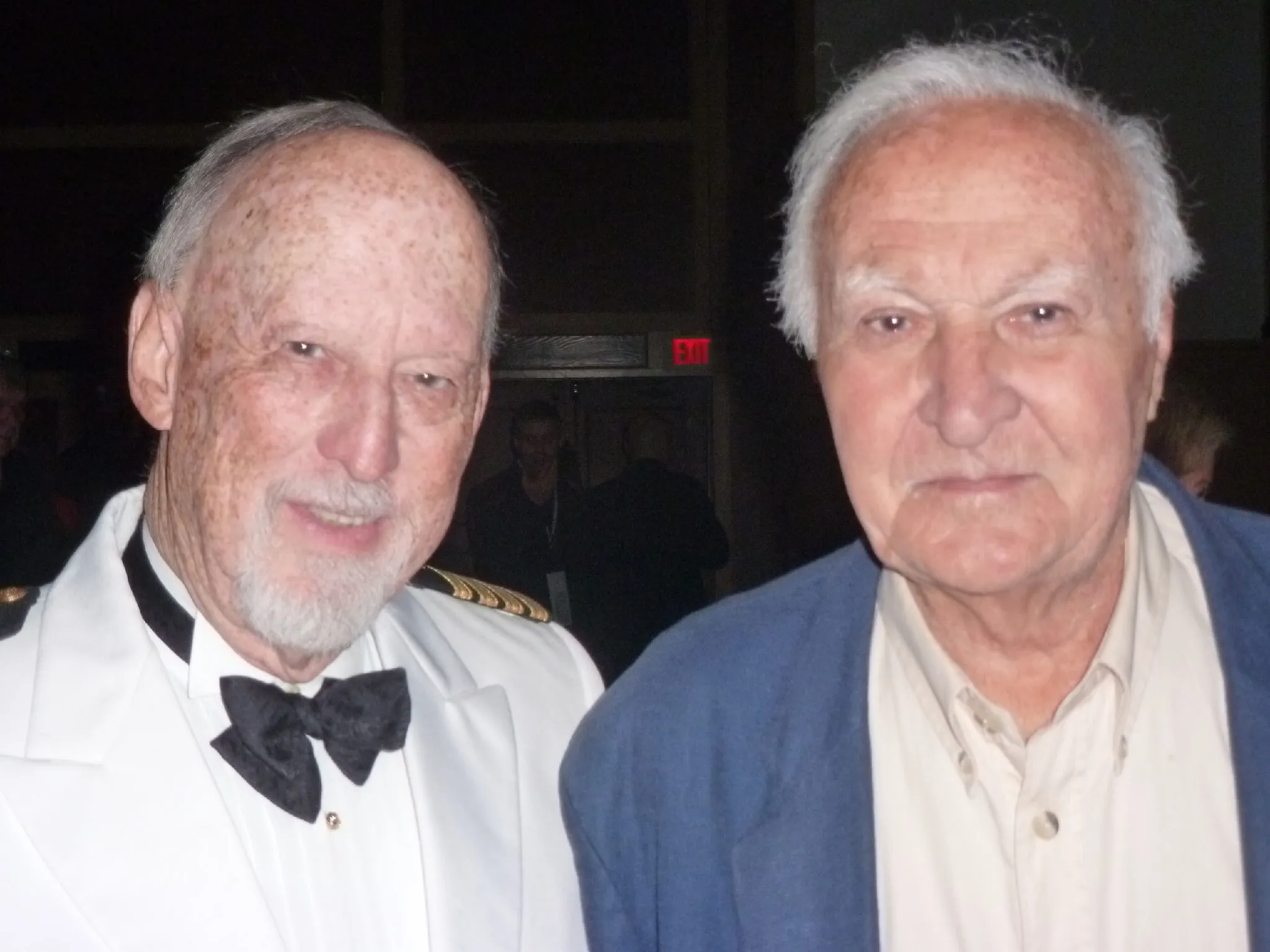 Portrait of two elderly men standing close together indoors; the man on the left is wearing a white naval uniform with a black bow tie, and the man on the right is dressed in a beige buttoned shirt and a blue jacket.