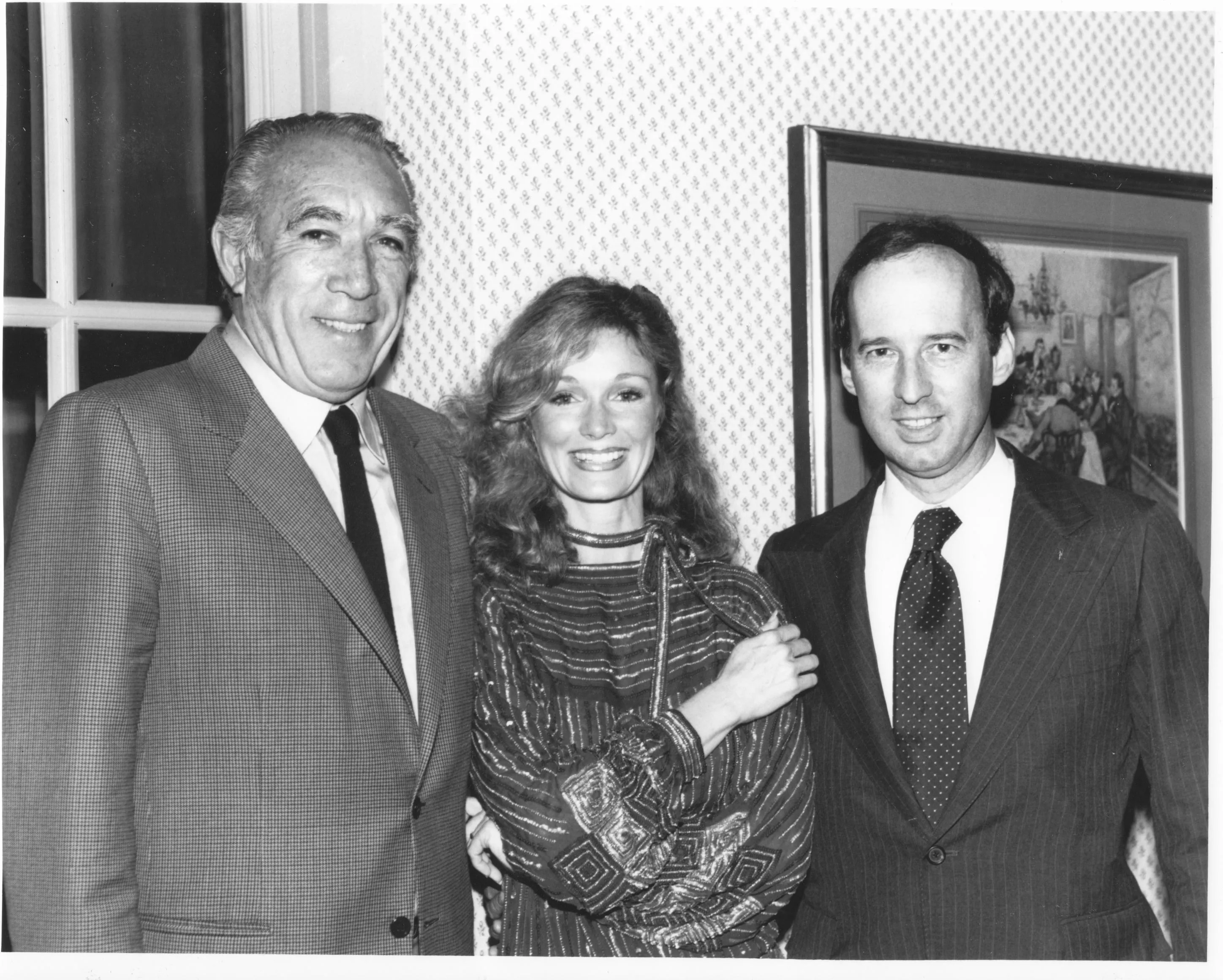 Black and white photo of three people standing indoors in front of a patterned wall and framed painting: an older man in a suit and tie on the left, a smiling woman with wavy hair in a patterned dress in the middle, and a younger man in a suit and tie on the right.