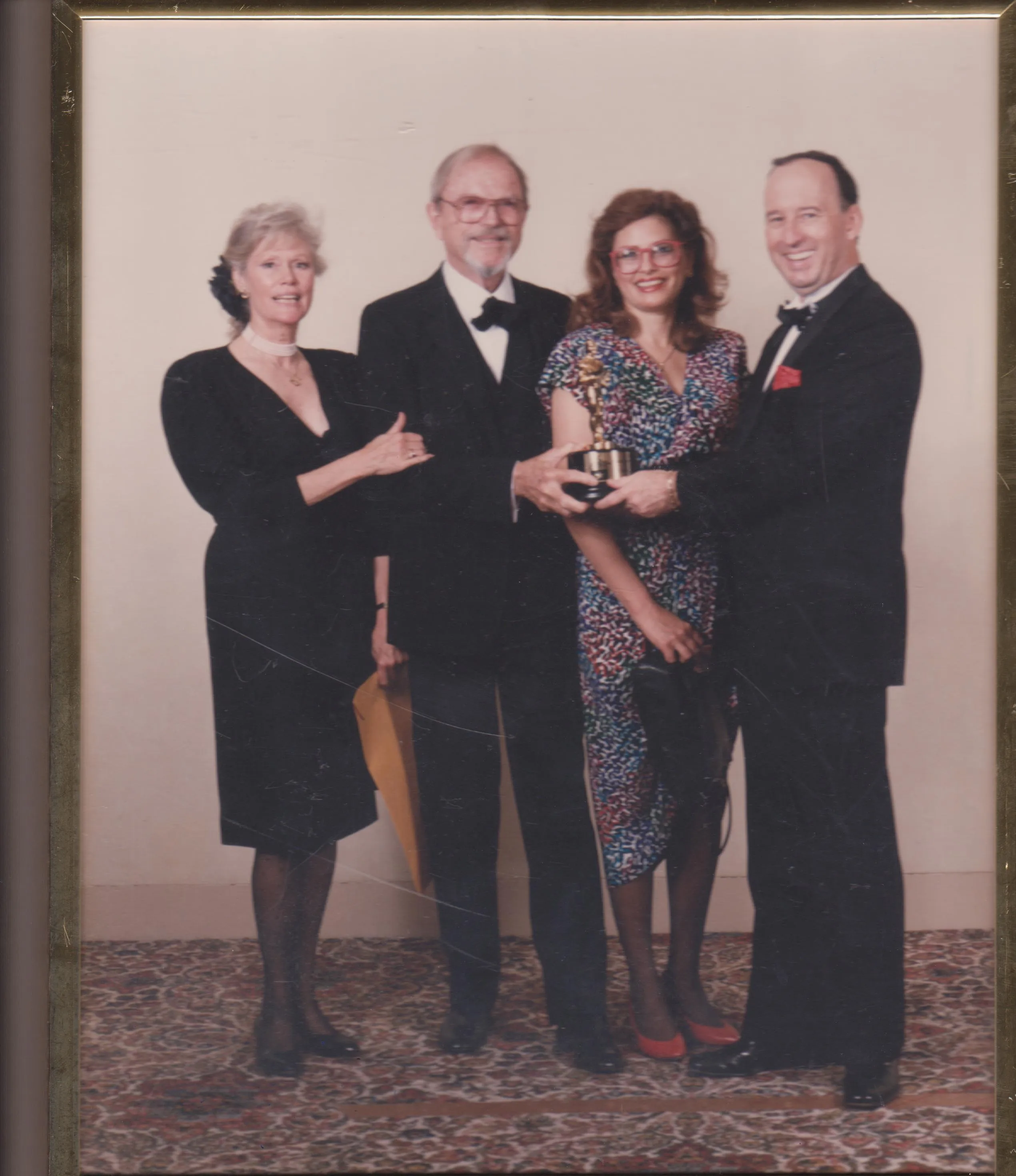 Four formally dressed people stand on a patterned carpet against a plain wall; two men in tuxedos and two women in dresses, one woman holds a golden statuette award with the man on the far right, everyone smiles.
