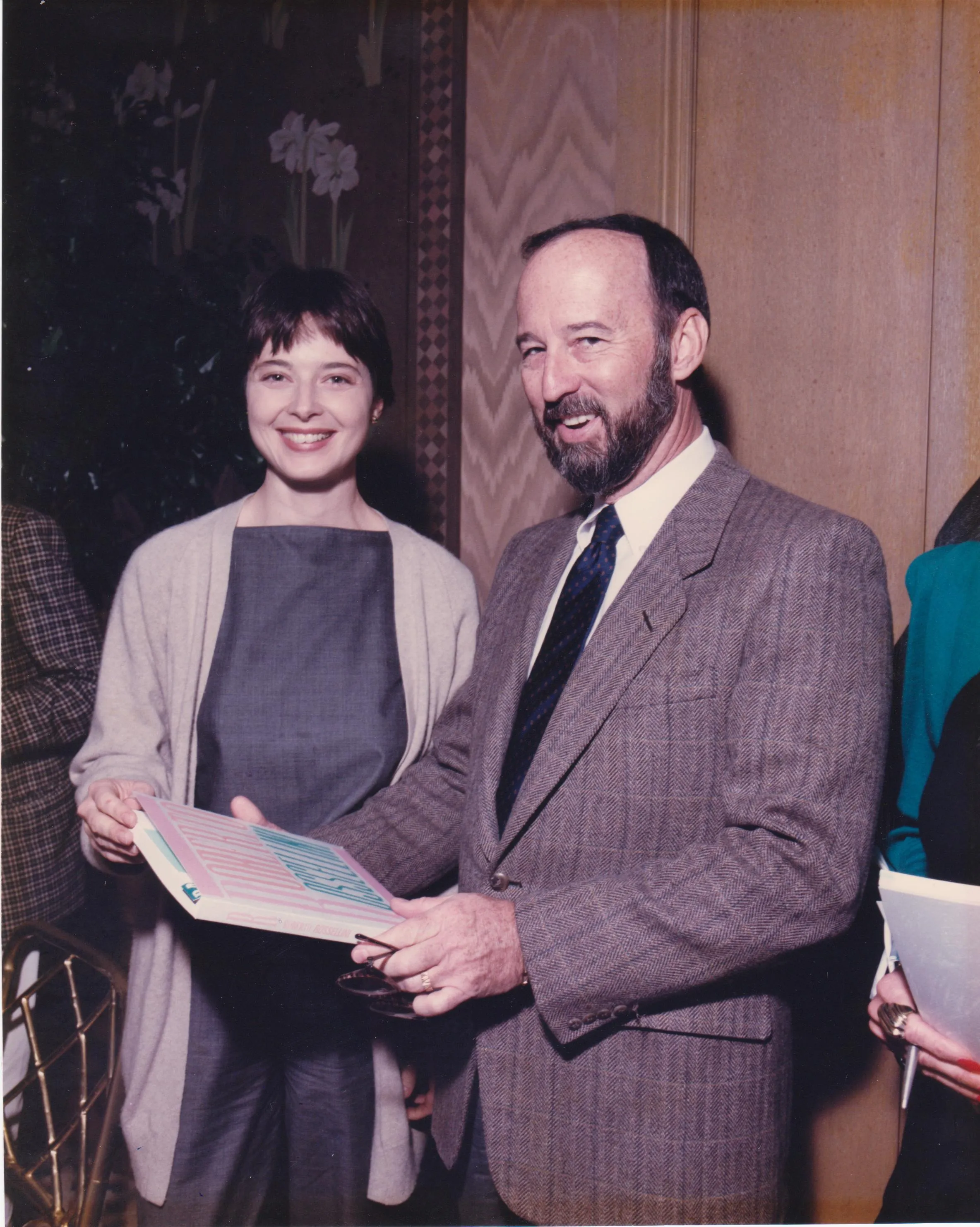 A smiling woman with short dark hair wearing a gray dress and light cardigan stands next to a bearded man in a brown checkered suit holding a book and eyeglasses in his hands in an indoor setting with patterned wallpaper and floral decor.