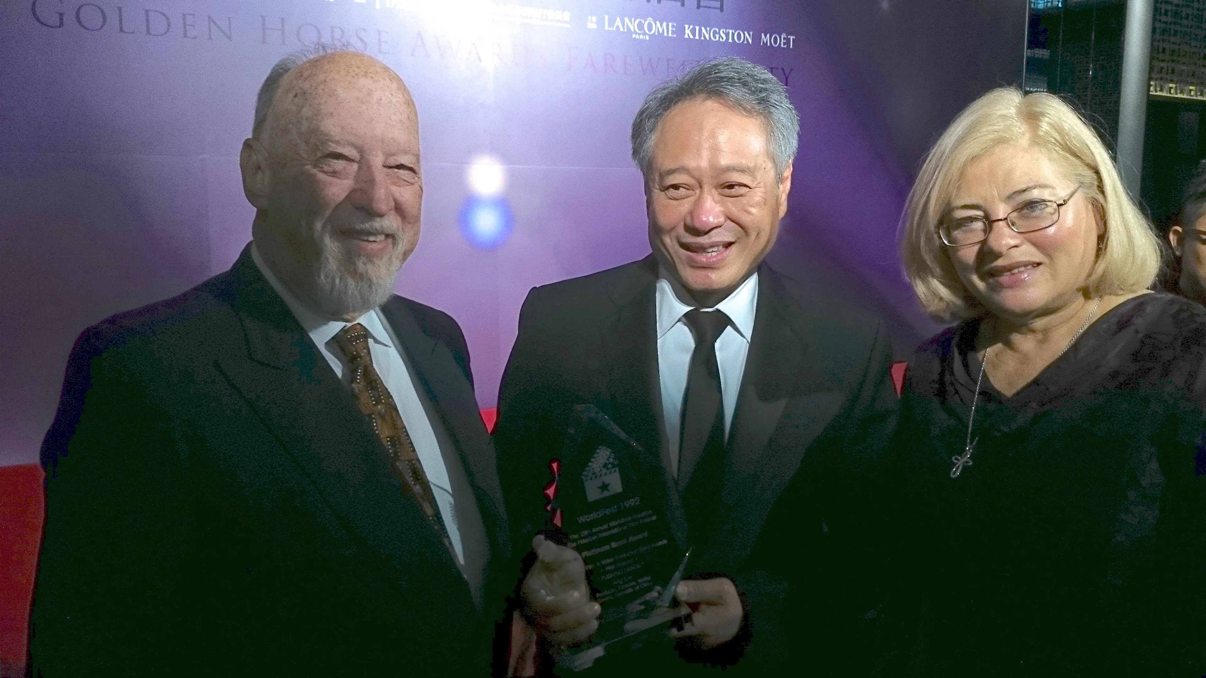 Three people dressed formally, an older man in a suit on the left, a middle-aged man in a black suit holding a glass award in the center, and a woman with blonde hair and glasses on the right, all smiling in front of a purple backdrop.