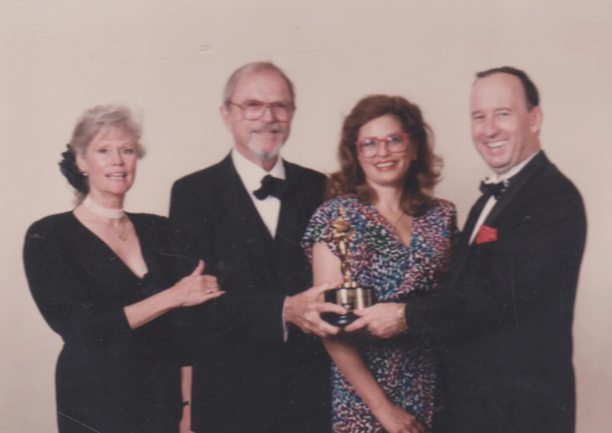 Four adults dressed formally, two men in tuxedos and two women in elegant dresses, holding a golden trophy together and smiling against a plain light background.