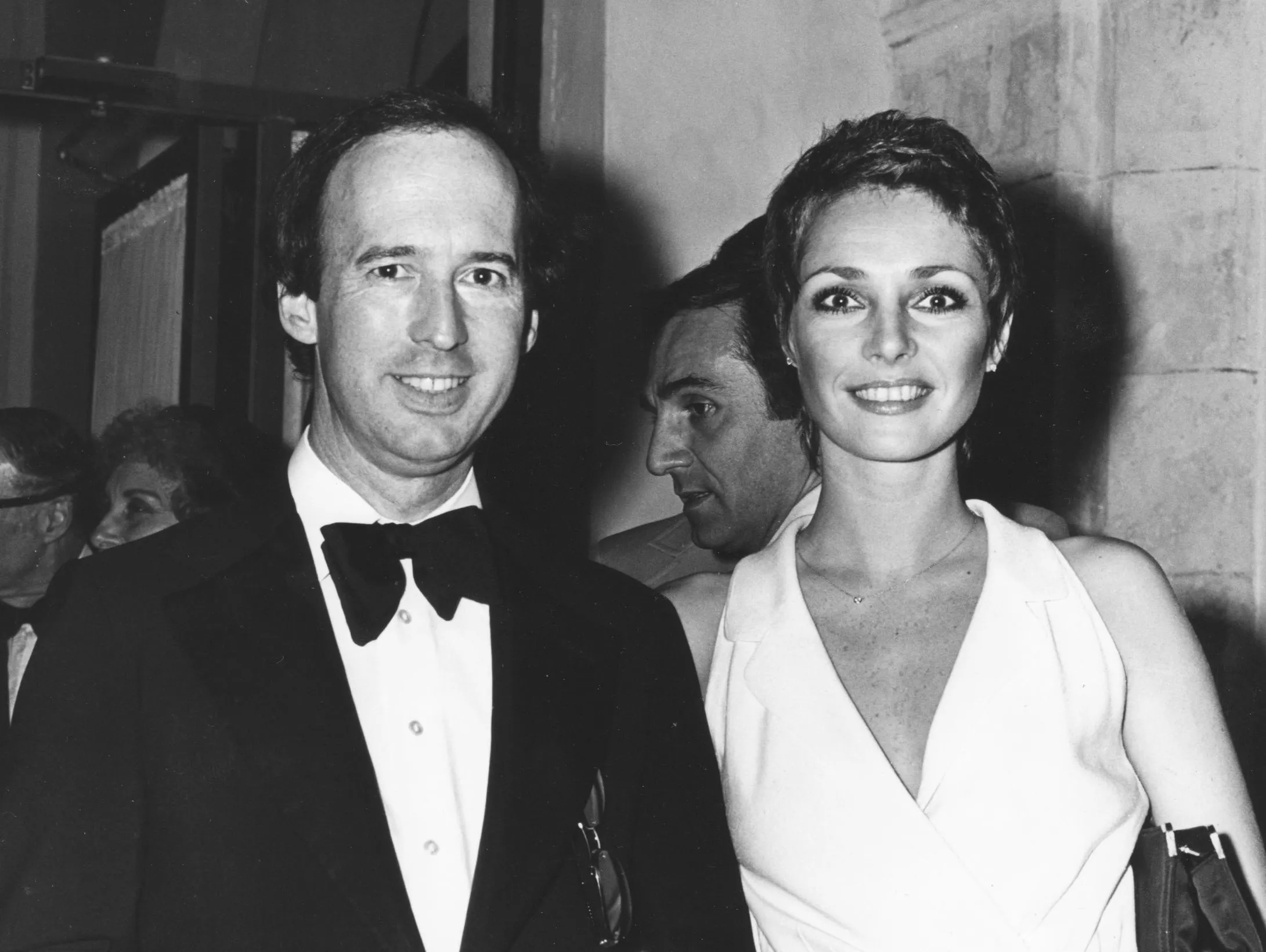 Black-and-white photo of a man in a tuxedo with a bow tie and a woman in a sleeveless white dress smiling at the camera in an indoor setting with people and stone walls in the background.