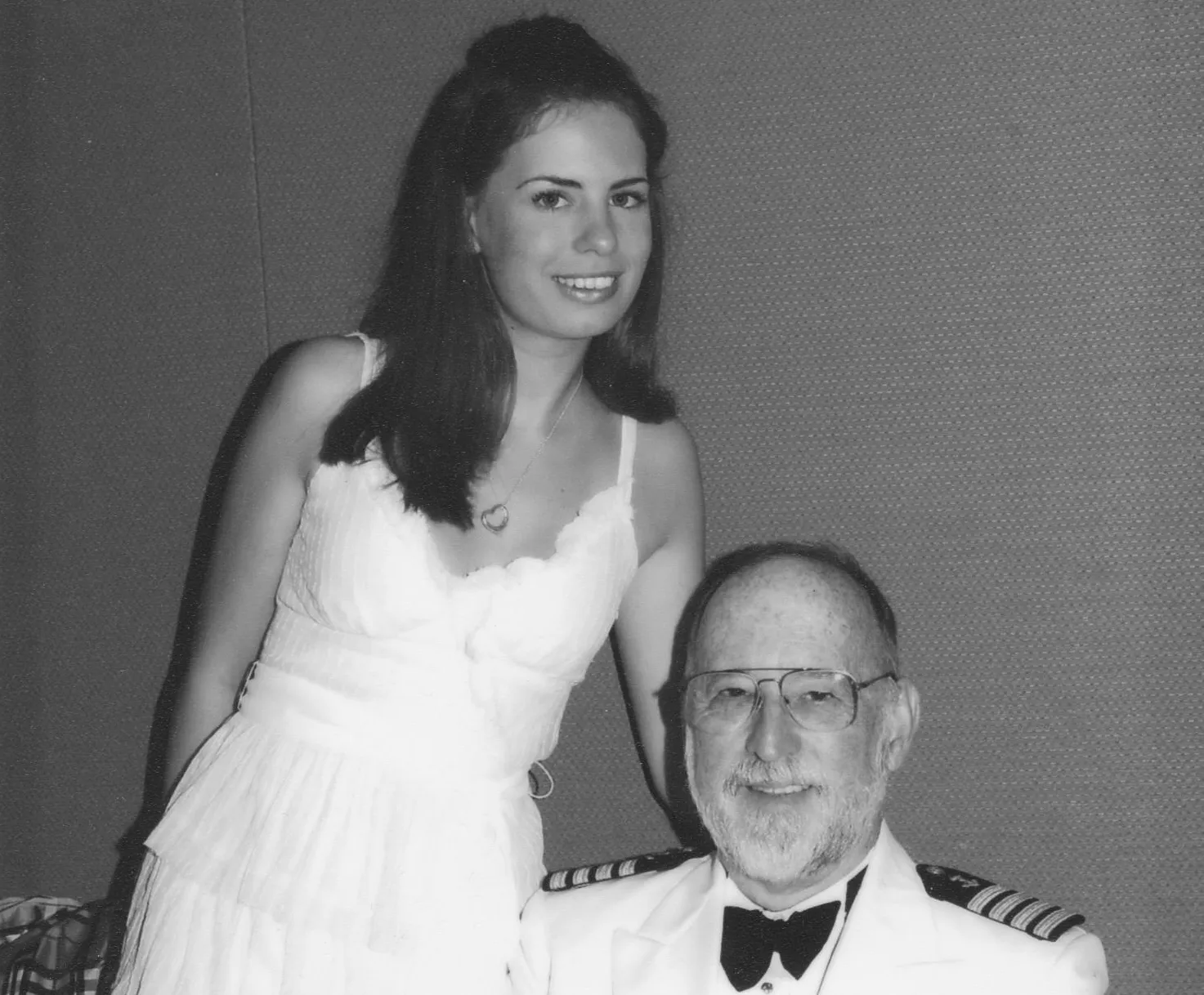 Black and white photo of a young woman in a sleeveless white dress with a heart necklace standing beside a seated older man wearing glasses, a white military dress uniform, and a bow tie.