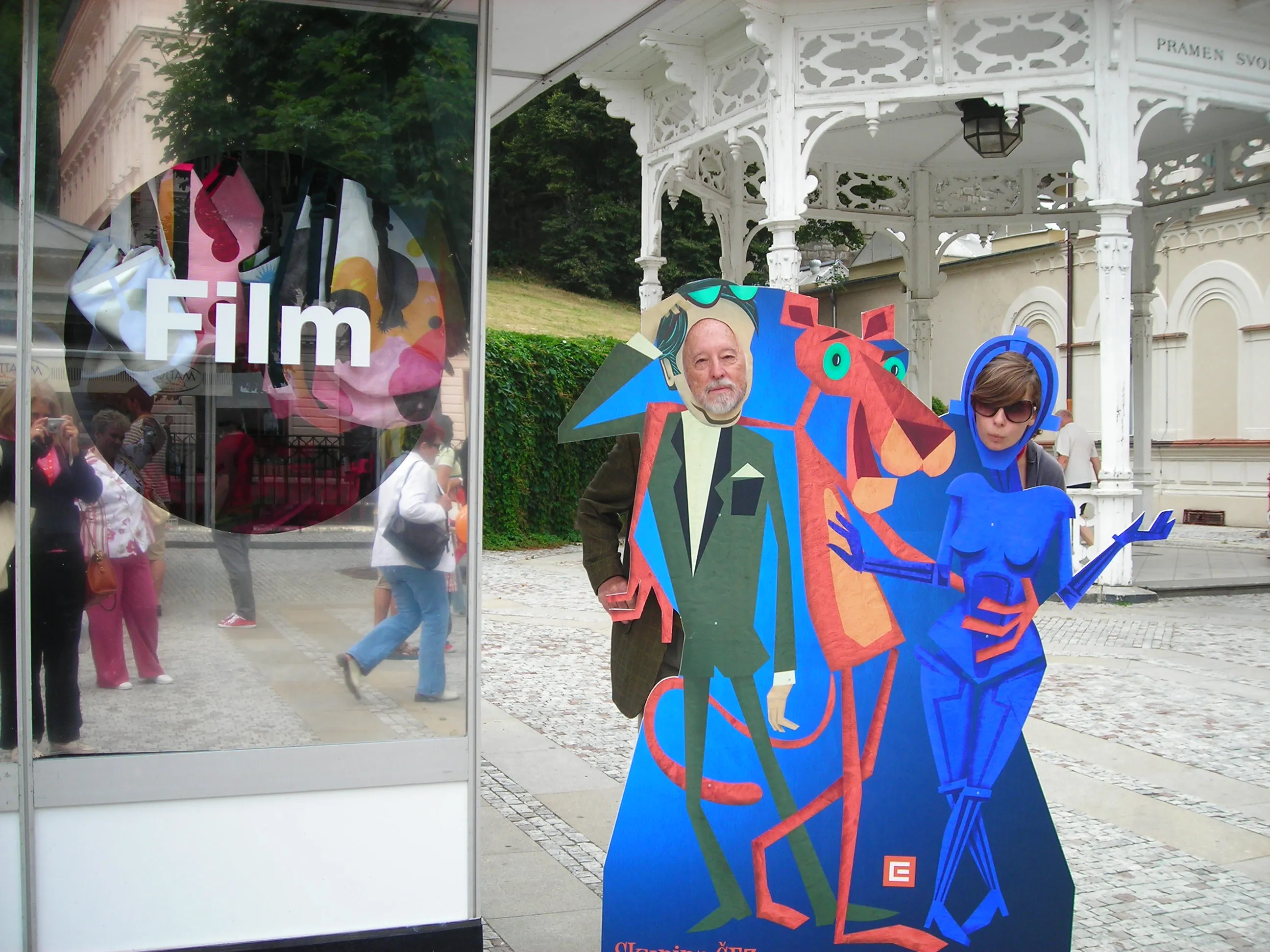 Two people posing with their faces showing through cutouts in a colorful cartoon standee featuring a man in a green suit, an orange panther-like animal, and a blue futuristic woman, set outdoors near a glass display with the word 'Film' and a white ornamental gazebo.